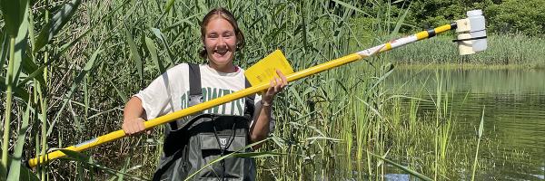 Lake Champlain Sea Grant Scholar Olivia Szumski, a Civil Engineering major, samples water in a University of Vermont stormwater pond as a summer intern with the campus Salt Mitigation Task Force. Student in waders stands in pond with long instrument.